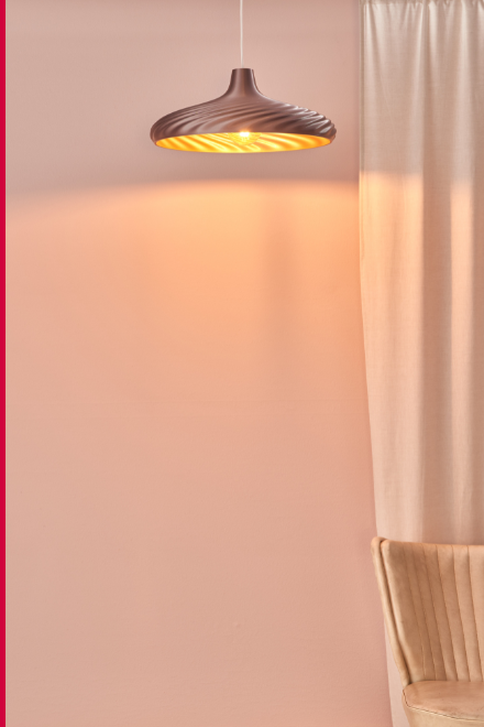 A hot-chocolate color pendant light with a spiral texture, casting a warm golden glow on a light pink wall with soft curtains nearby.